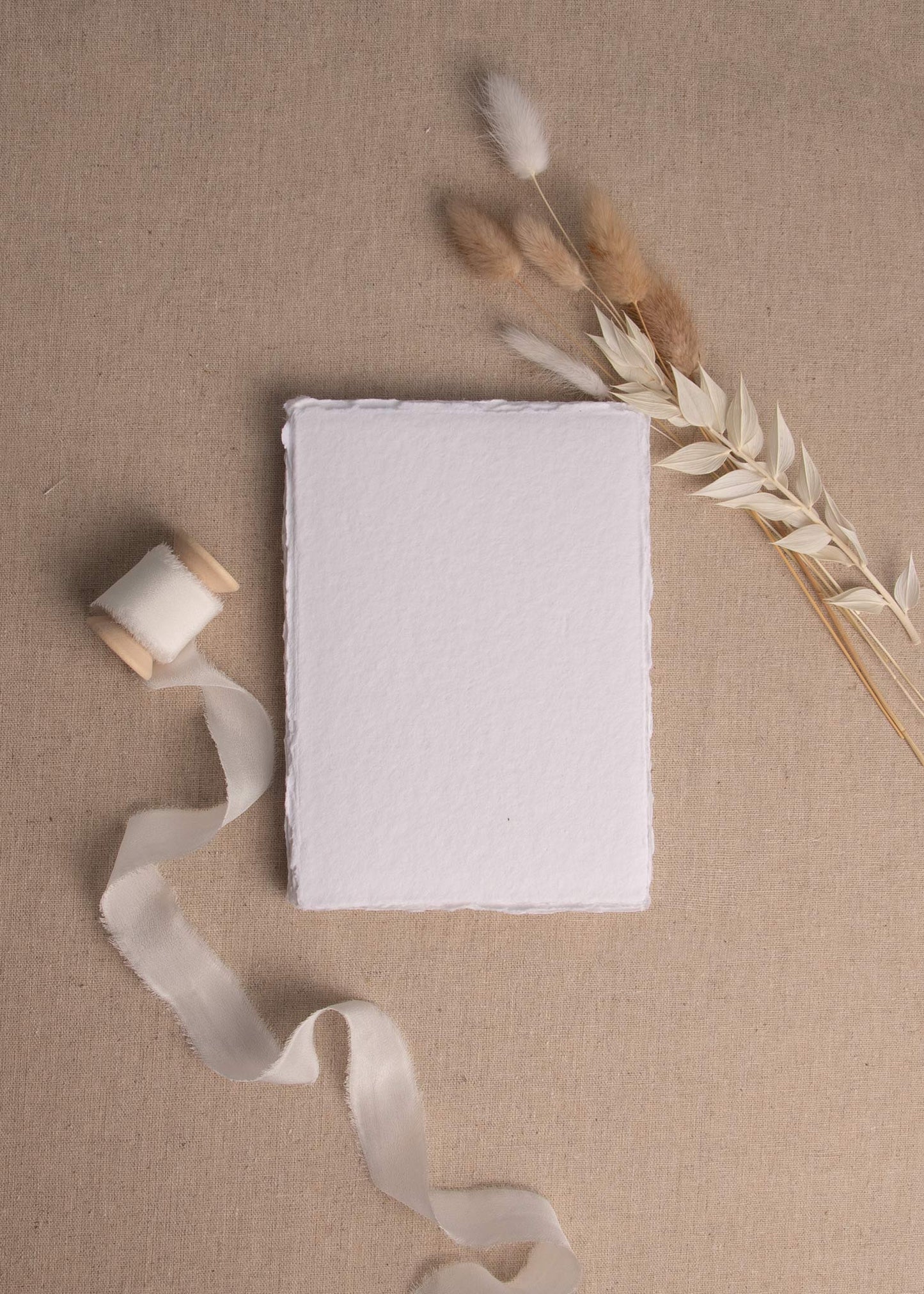 White Handmade paper with deckle edge on linen background surrounded by dried flowers and white silk ribbon spool