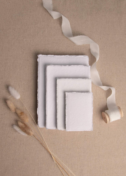 Pile of various sizes of white handmade paper with deckle edge surrounded by dried flowers and white silk ribbon spool
