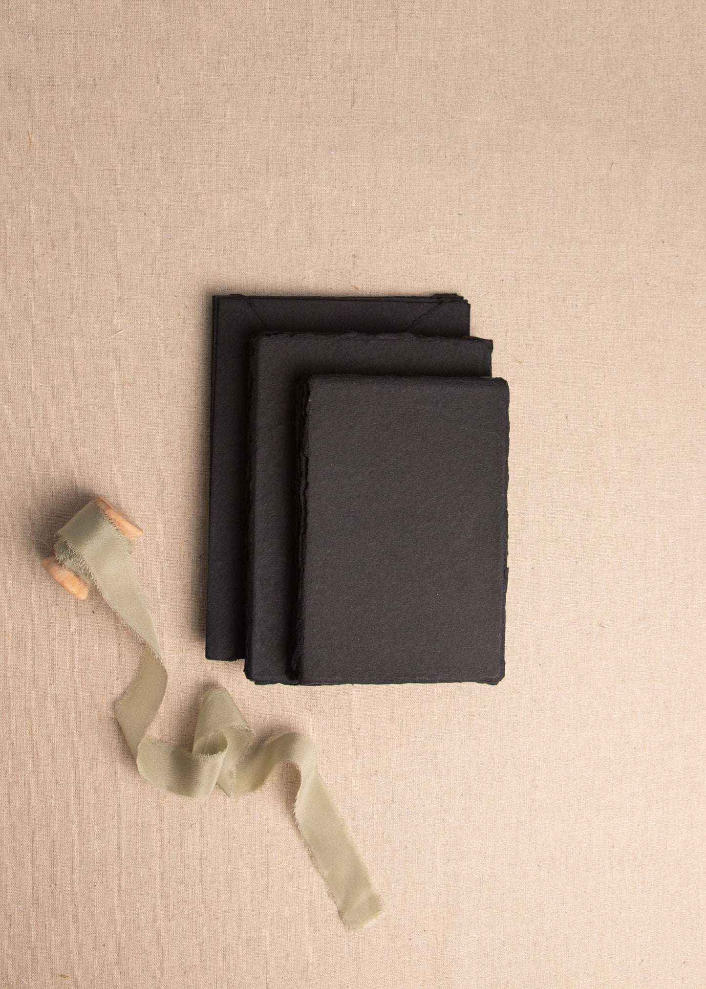 Stack of 5x7 inch Black Handmade paper and envelope with deckle edge surrounded by sage green ribbon spool