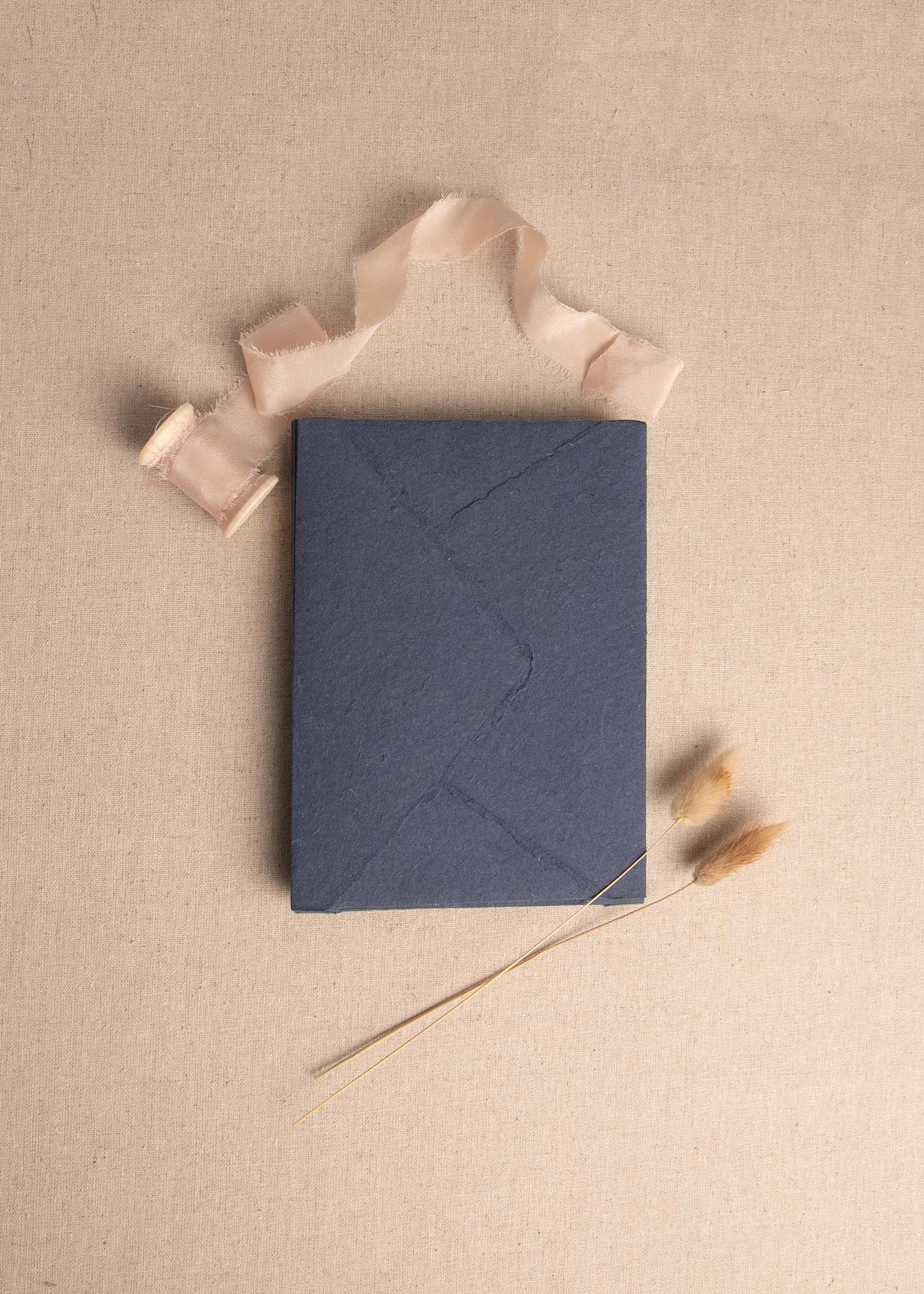 Singular 5x7 Dark Blue Handmade paper envelope with deckle edge surrounded by champagne colour silk ribbon spool and bunny tail dried flowers