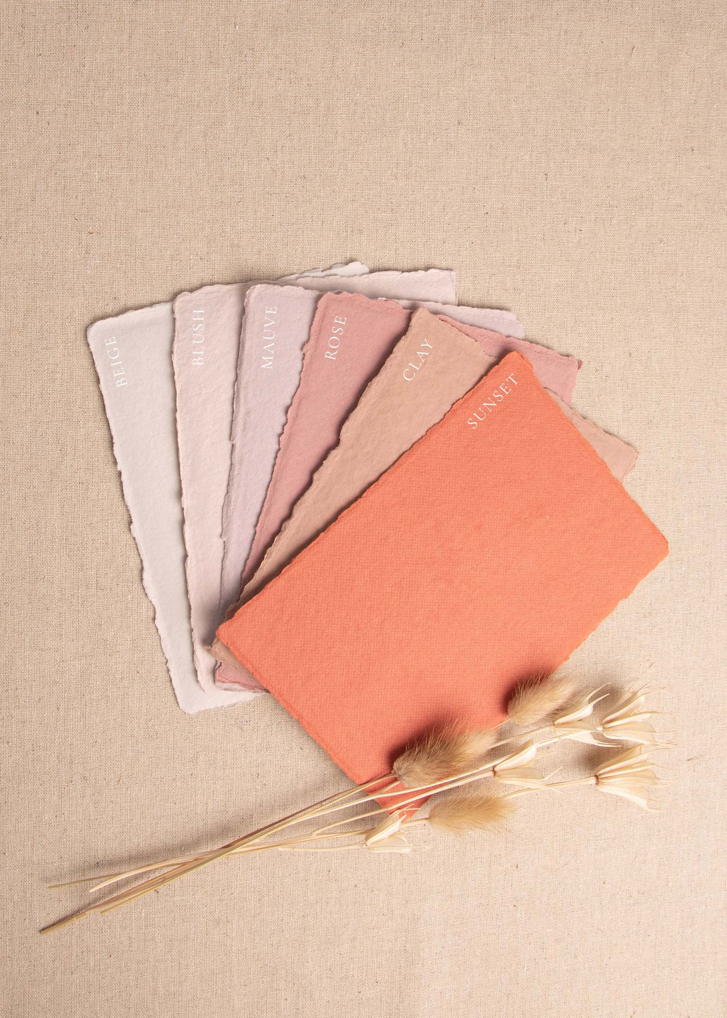 Fanned pile of Beige, Blush, Mauve, Rose, Clay, Sunset handmade paper envelopes with deckle edge surrounded by dried flowers