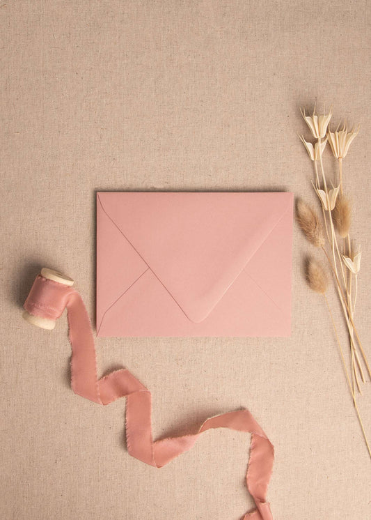Elevated view of blush dusky pink wedding envelopes surrounded by silk ribbon spool and dried flowers