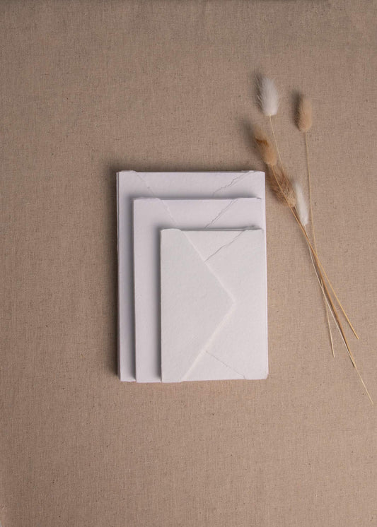 Pile of various sizes of white handmade paper envelopes with deckle edge surrounded by dried flowers on linen background