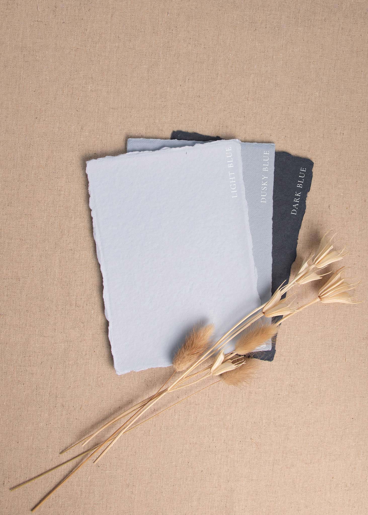 Fanned pile of Light blue, Dusky blue, Dark Blue handmade paper envelopes with deckle edge surrounded by dried flowers