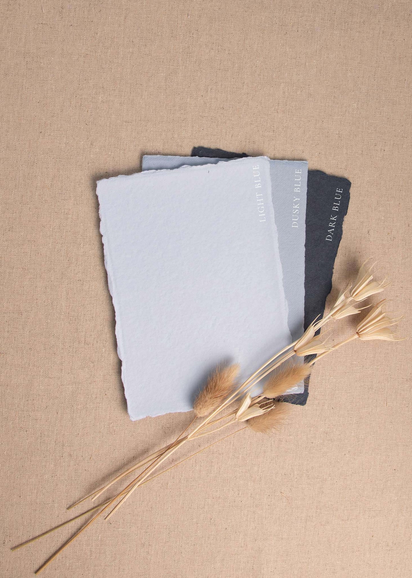 Fanned pile of Light blue, Dusky blue, Dark Blue handmade paper sheets with deckle edge surrounded by dried flowers