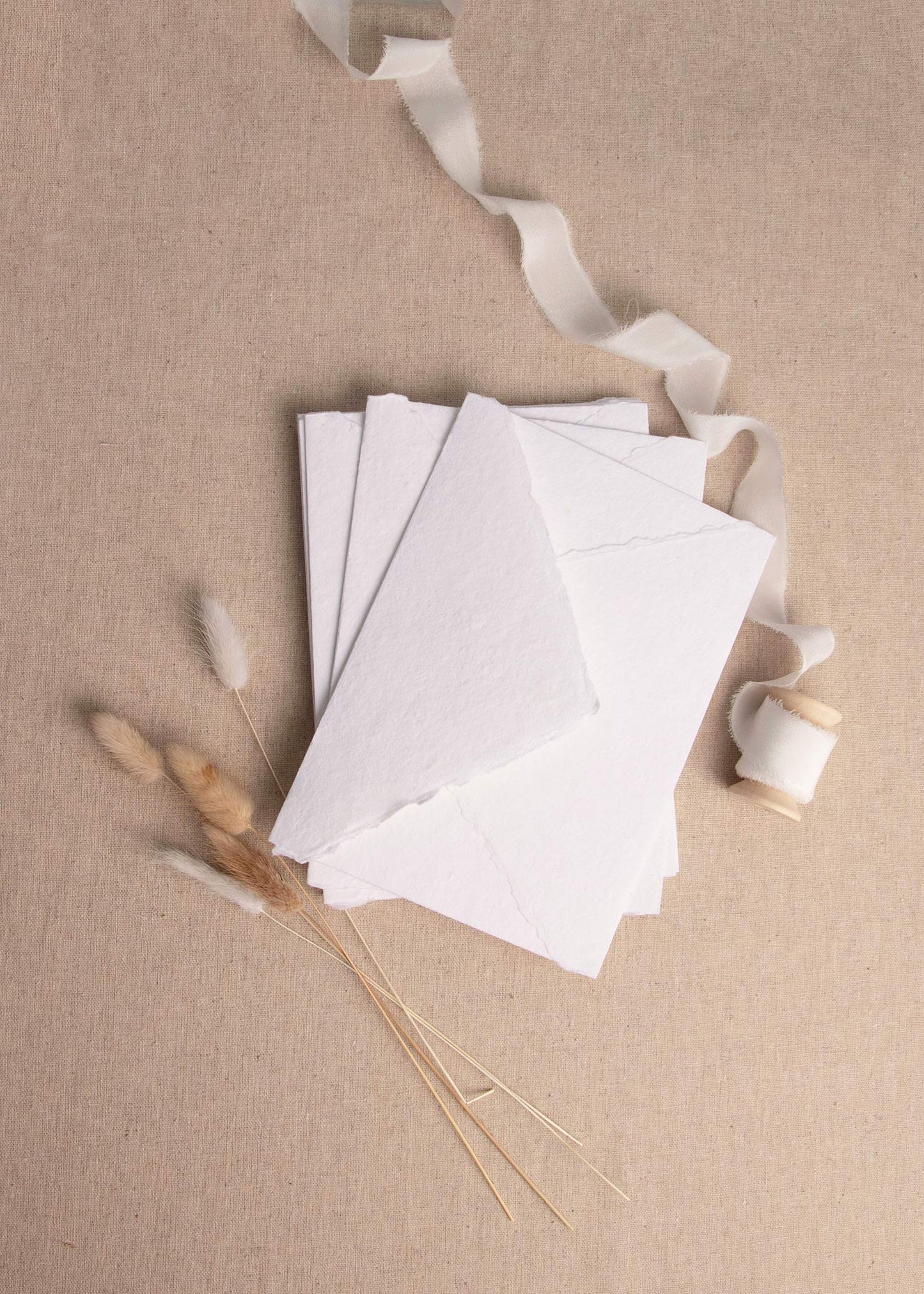 Fanned pile of White handmade paper with deckle edge surrounded by dried flowers and white silk ribbon spool