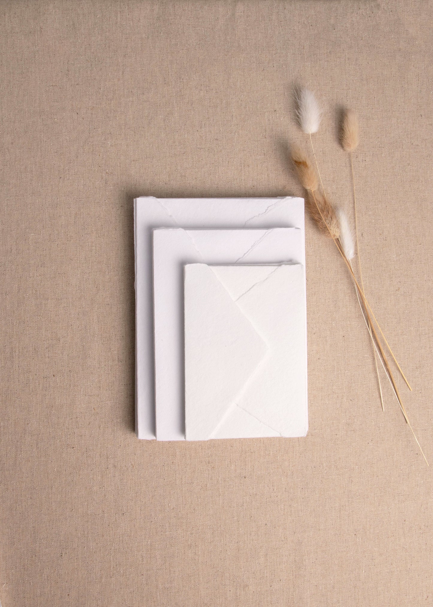 Pile of various sizes of White handmade paper and envelopes with       4. deckle edge on linen background with dried flowers